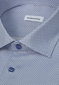 Chemise habillée à col avec motif géométrique bleu, deux boutons bleus visibles et étiquette "seidensticker" à l'intérieur du col.