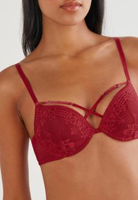Red lace bra with a floral pattern, featuring crisscross straps, underwire support, and a smooth textured finish.