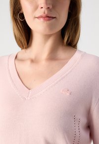 Šviesiai rožinė V formos megztinis, pagamintas iš minkštos trikotažo medžiagos, su raštuotais kraštais ir nedideliu siuvinėtu logotipu krūtinėje bei subtiliu dekoratyviniu siuvimu.