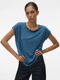 Femeie cu părul dat pe spate, purtând un top teal cu mânecă scurtă și blugi închiși la culoare, cu mâinile strângând marginea tricoului.