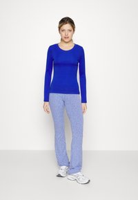 Blaues, geripptes Langarmshirt kombiniert mit hellblauen, gemusterten Schlaghosen. Das Modell trägt graue Sportschuhe; schlichter weißer Hintergrund.