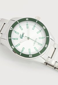 Ασημένιο μεταλλικό ρολόι χειρός με πράσινο στεφάνι και δείκτες, λευκό καντράν, λογότυπο Lacoste και πράσινο έμβλημα κροκοδείλου σε λευκό φόντο.