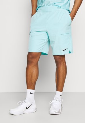 Man draagt lichtblauwe sportshorts, witte Nike-sokken en witte Nike-sneakers, staat met handen in de zakken.