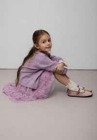 Dziewczyna siedzi na jasnoszarej powierzchni, ubrana w lawendowy sweter, warstwową różową spódnicę z kwiatowymi wzorami, białe skarpetki i różowe buty na paski.