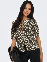 Femme aux longs cheveux bruns portant un chemisier beige à imprimé léopard noir avec des manches bouffantes, tenant un sac à main noir froncé à son côté.