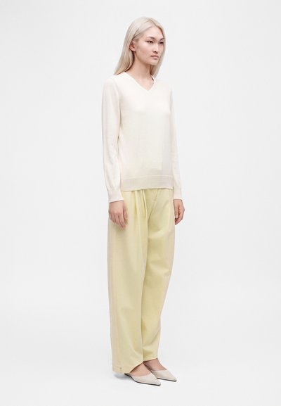 Νεαρή γυναίκα με μακριά ξανθά μαλλιά, που φοράει κρεμ πουλόβερ με V λαιμόκοψη, ανοιχτό κίτρινο παντελόνι με φαρδιά μπατζάκια και κρεμ αιχμηρά παπούτσια, στέκεται μπροστά σε ένα λευκό φόντο.