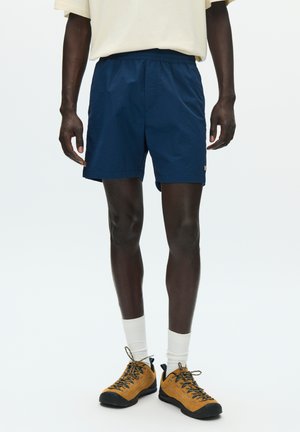 Marineblå atletikshorts med elastisk talje, der har et lille logo, kombineret med beige sko og hvide mellemlange sokker.