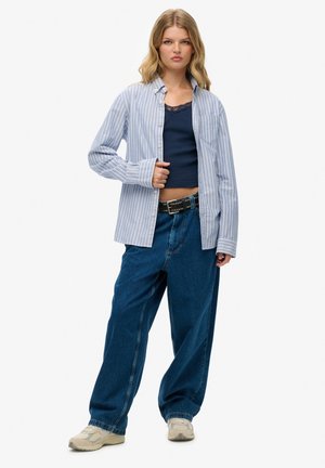 Jonge vrouw in losse blauwe spijkerbroek, zwarte riem, marineblauw topje met kant, opengeknoopte blauw-wit gestreepte blouse en beige sneakers, staand.