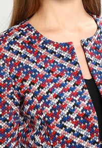 Lähikuva henkilöstä, jolla on päällään monivärinen kudottu takki, jossa on punaisia, sinisiä, valkoisia ja mustia geometrisiä kuvioita, sekä pyöreä kaulus.