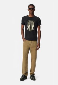 Camiseta negra de manga corta con un estampado gráfico, combinada con pantalones kaki de pierna recta y zapatos de plataforma negros. Modelo con gafas de sol.