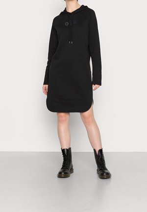 Vestito nero con cappuccio realizzato in morbido tessuto, caratterizzato da maniche lunghe, orlo arrotondato e spacchi laterali. Abbinato a stivali da combattimento neri con lacci.
