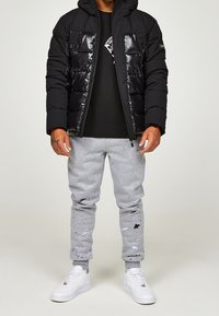 Svart vadderad jacka med glansig yta, grå sweatpants med färgfläckar och vita sneakers. Huva ingår på jackan.