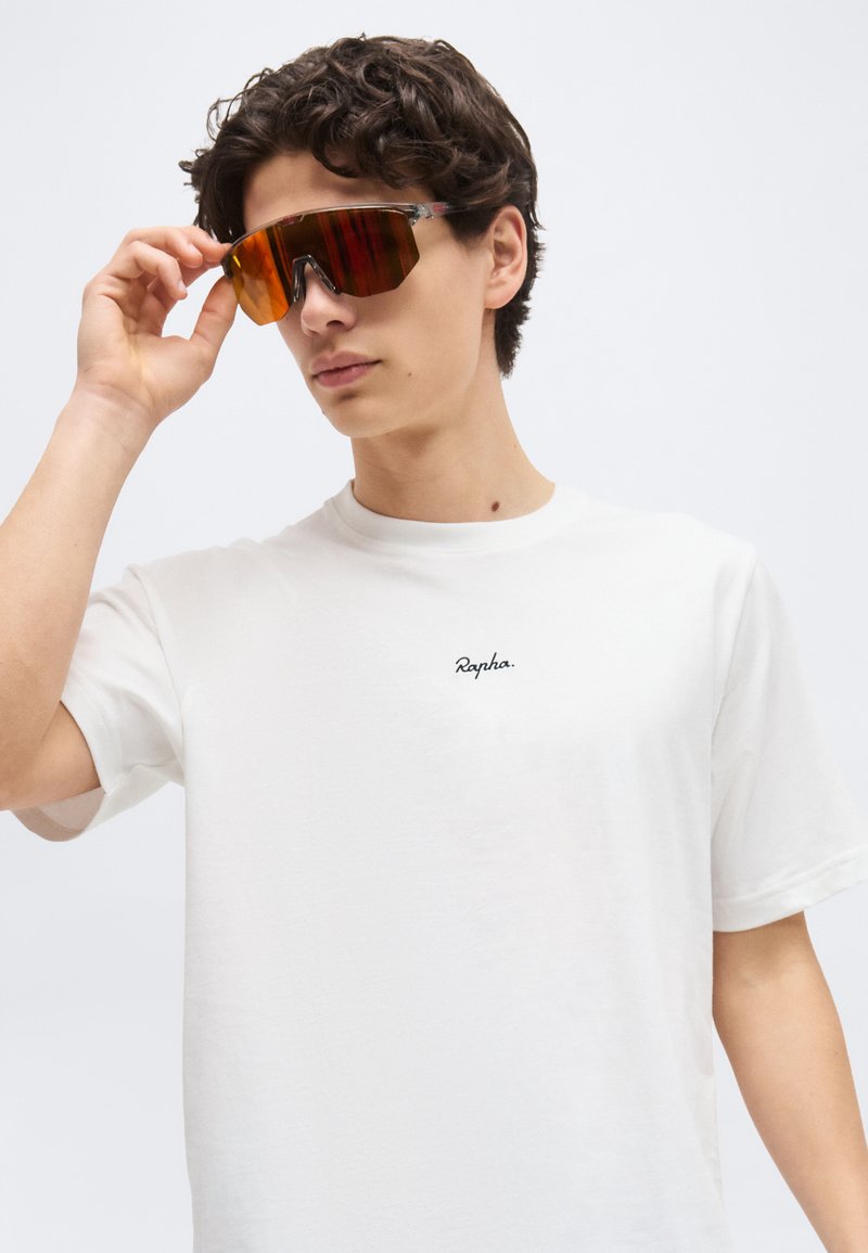 Young man adjusting reflective orange sports sunglasses, wearing a white Rapha t-shirt against a plain light background.