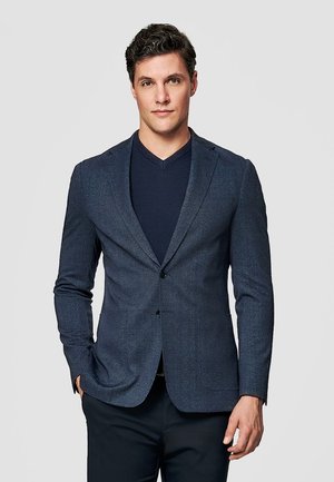 Młody mężczyzna z krótkimi ciemnymi włosami, ubrany w granatową marynarkę na ciemnej koszulce z dekoltem w serek, stoi na tle jednolitego jasnoszarego tła.