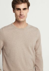 Beige, rundhalsad tröja gjord av mjukt material. Har långa ärmar och en enkel design. Texturen är slät och har en lätt melerad stil.