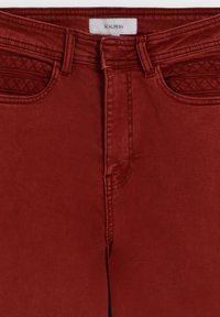 Jeans roșii cu detalii matlasate pe buzunarele frontale și buclele pentru curea, eticheta vizibilă a brandului Scalpers în căptușeala taliei.