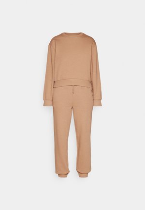 Sweat-shirt à manches longues couleur beige et pantalon de jogging assorti avec des poignets élastiques, présenté sur un fond gris clair uni.