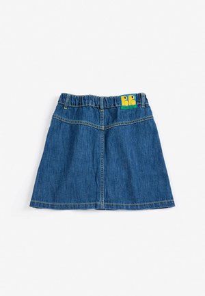 Kind mit kurzen blonden Haaren, das ein grünes T-Shirt mit gelbem Text, einen Jeansrock mit Knöpfen, gemusterte Socken und braune Schuhe trägt, steht vor einem einfarbigen Hintergrund.