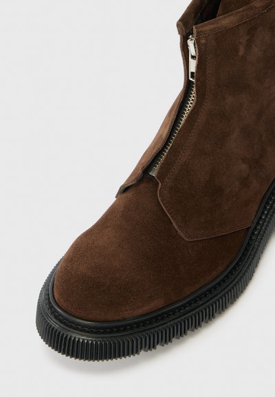 Botte de cheville en suède marron avec une fermeture éclair sur le côté et une semelle en caoutchouc noir texturé. Elle présente un dessus lisse, un bout rond et une ouverture inclinée.