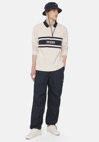 Beżowa koszulka polo z długim rękawem w granatowe paski i logo "DICKIES", zestawiona z granatowymi spodniami cargo i białymi sneakersami, dopełniona granatowym kapeluszem typu bucket.