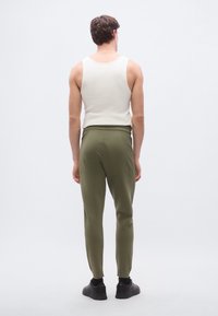 Pantalones deportivos para hombre en verde oliva con textura acanalada y piernas ajustadas, combinados con una camiseta sin mangas blanca, vistos desde atrás. Zapatillas negras.