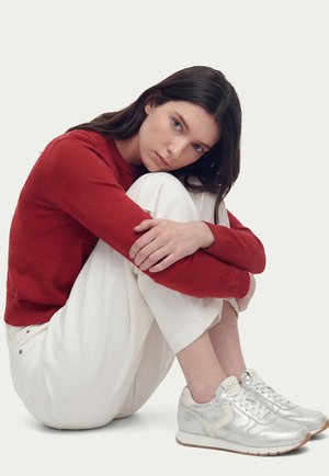 Giovane donna con capelli lunghi e scuri seduta abbracciando le ginocchia, indossa un maglione rosso, pantaloni bianchi e scarpe da ginnastica argentate contro uno sfondo semplice.