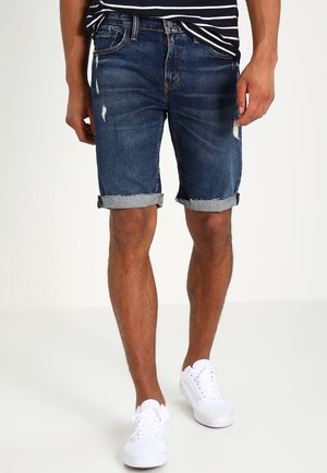 Pantalones cortos de mezclilla en azul oscuro con desgastes, dobladillos enrollados y un ajuste slim. Llevados con una camiseta de rayas en azul marino y blanco y zapatillas blancas.