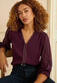 Bordo blūze ar V veida kakla izgriezumu un dekoratīvām pogu akcentiem. Iespējo ruffles pie piedurknēm un audumu ar tekstūru un delikātu šuvumu.