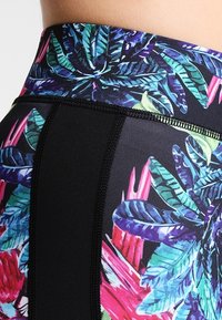 Leggings noirs taille haute avec un motif floral coloré, comprenant des feuilles vertes et des accents roses, fabriqués à partir d'un matériau extensible et texturé.