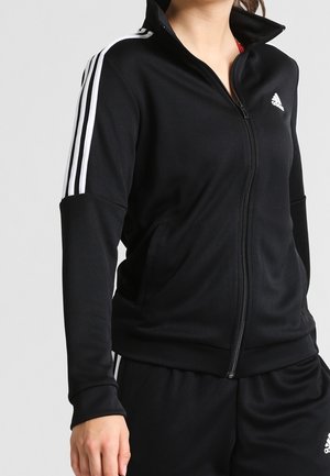 Woman wearing a black Adidas tracksuit with white stripes on sleeves and Adidas logo on chest, arms relaxed by sides.