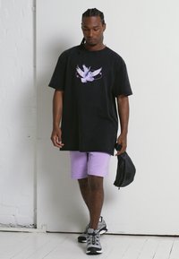 Camiseta negra oversized con un gráfico de un pájaro blanco y morado, combinada con pantalones cortos morados claros y zapatillas deportivas grises. Lleva una bolsa negra en la mano.