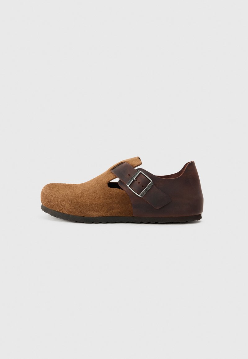 Braune Wildleder-Clogs mit einer abgerundeten Spitze, seitlichen Ausschnitten und einem dunkelbraunen Lederriemen mit einer silbernen Schnalle. Strukturiertes Obermaterial und Gummisohle.