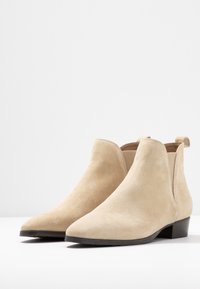 Beige mokkasiini nilkkurit, joissa on teksturoitu pinta, kärki, matala korko ja joustavat sivupaneelit, somat käyttömukavuuteen. Tasainen sisävuori.