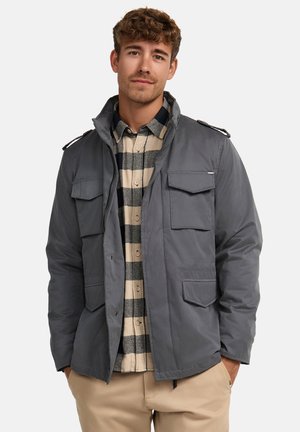 Chaqueta gris aislante con múltiples bolsillos frontales y cuello alto. Llevada sobre una camisa a cuadros en tonos beige y azul marino. Ajuste y diseño casual.