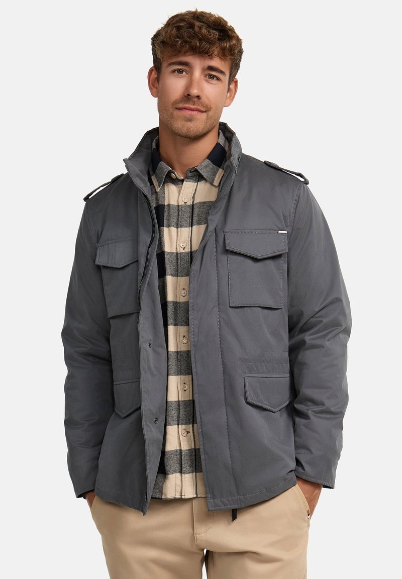 Veste grise isolée avec plusieurs poches à l'avant et un col montant. Portée par-dessus une chemise à carreaux beige et marine. Coupe et design décontractés.