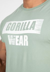Groene katoenen t-shirt met "GORILLA" in vetgedrukte witte strepen en "WEAR" in solide wit, korte mouwen en een ronde hals.