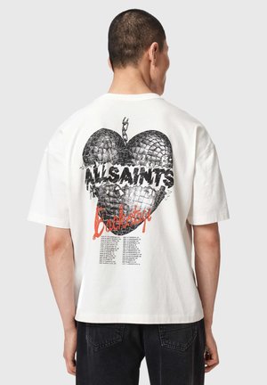 Mężczyzna noszący białą koszulkę z czarną grafiką złamanego serca, tekstem „ALLSAINTS Backstore” oraz listą nazw miast na plecach.