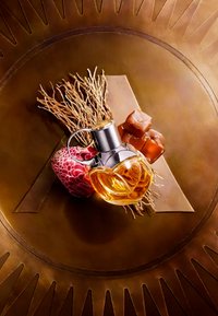 Transparante glazen parfumfles met een bloemmotief, omringd door karamelblokjes, plantwortels en een gestructureerd roze object op een bronzen achtergrond.