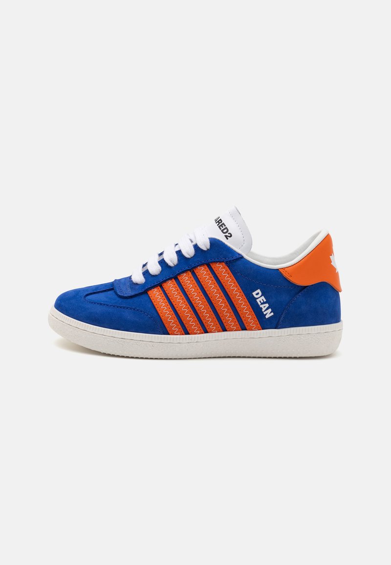 Zapatilla de ante azul con tres franjas naranjas prominentes, cordones blancos y suela de goma blanca. Presenta un acento en el talón en naranja con la marca.