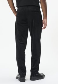 Homme portant un pantalon noir sur mesure et des chaussures en cuir noires, se tenant les mains dans les poches sur fond blanc.