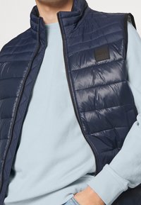 Marinblå quiltad väst med hög krage och dragkedja, med en logotyp-patch; bärs över en ljusblå sweatshirt. Slät, glänsande textur.