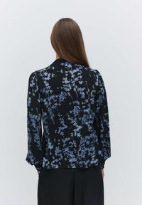 Svart blus med blått blommönster, långa ärmar, mjukt tyg, rund halsringning och rynkade detaljer vid axlarna. Knäppning i ryggen.