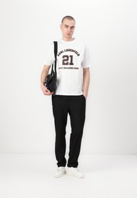 Hvit bomull T-skjorte med mørk tekst og nummer "21," kombinert med svarte bukser og hvite sneakers. Modellen holder en liten svart veske.