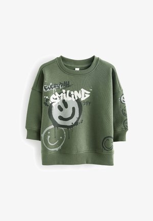 Sweat-shirt vert olive avec poignets et ourlet côtelés, arborant de grands et petits visages souriants peints ainsi que le texte "EVERYDAY SMILING 24/7" sur le devant et la manche.