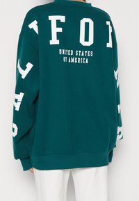 Sudadera de gran tamaño en color azul verdoso con letras blancas en la parte posterior que dicen "FOI" y "ESTADOS UNIDOS DE AMÉRICA." Mangas largas con letras adicionales.