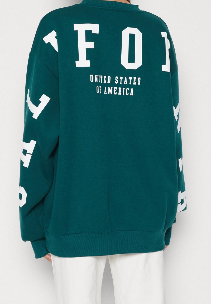 Sudadera de gran tamaño en color azul verdoso con letras blancas en la parte posterior que dicen "FOI" y "ESTADOS UNIDOS DE AMÉRICA." Mangas largas con letras adicionales.