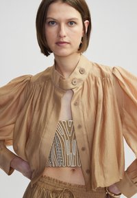 Lichtbeige cropped blouse met pofmouwen, knoopsluiting en een hoge kraag. Daaronder een gebloemde beige bandtop met versieringen.