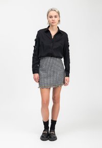 Schwarzes Button-Up-Hemd mit strukturierten Ärmeln, kombiniert mit einem schwarz-weißen Hahnentritt-Minirock mit fransigem Saum. Schwarze Plateauschuhe.