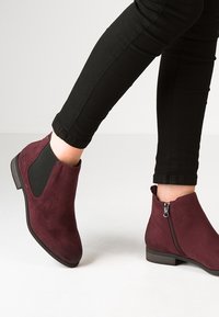 Piernas en pantalones negros usando botines de ante burdeos con cremallera lateral y panel elástico sobre un fondo claro.