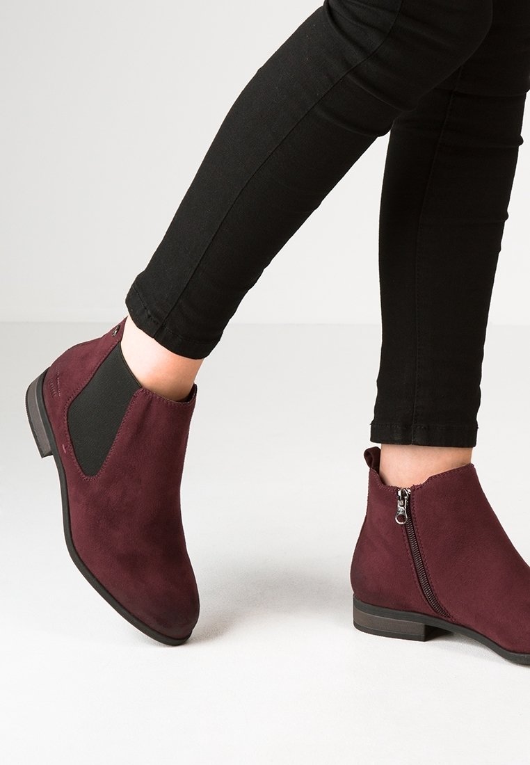 Gambe in pantaloni neri che indossano stivaletti alla caviglia in suede bordeaux con cerniera laterale e pannello elastico su uno sfondo chiaro.